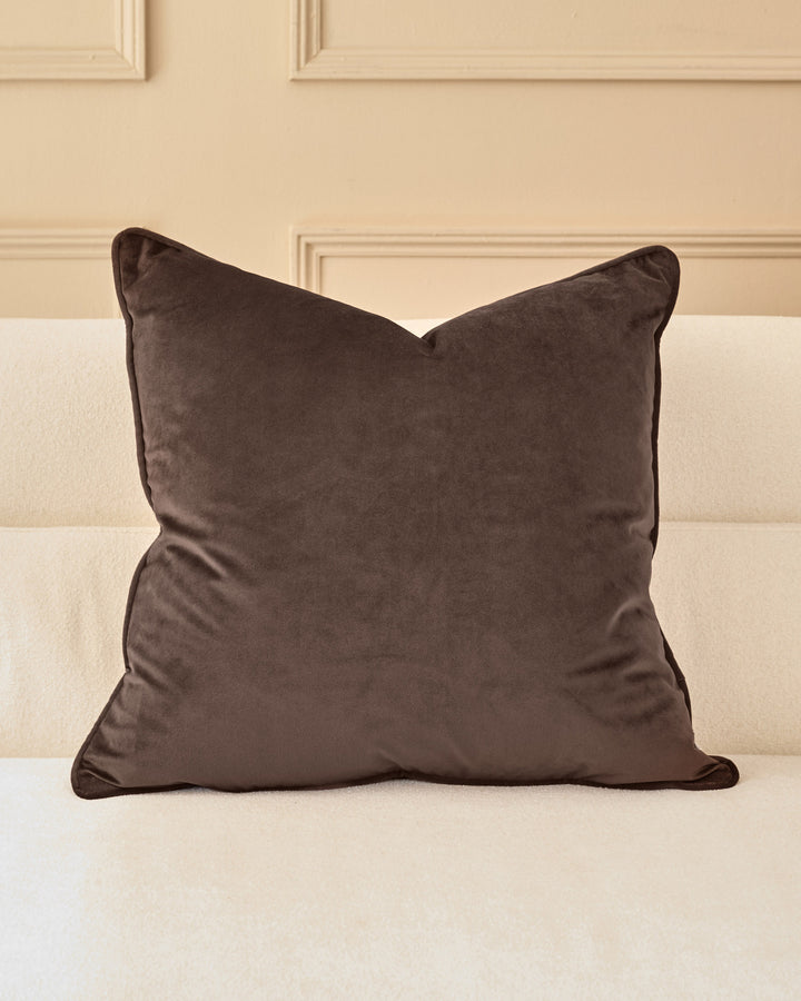 Large chocolate velvet cushion with piping detail, displayed on a cream upholstered bed against a beige paneled wall.