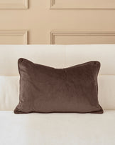 Brown velvet rectangular cushion with piping detail, displayed on a cream upholstered headboard against a beige paneled wall.