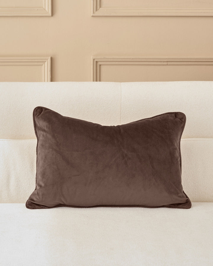 Brown velvet rectangular cushion with piping detail, displayed on a cream upholstered headboard against a beige paneled wall.