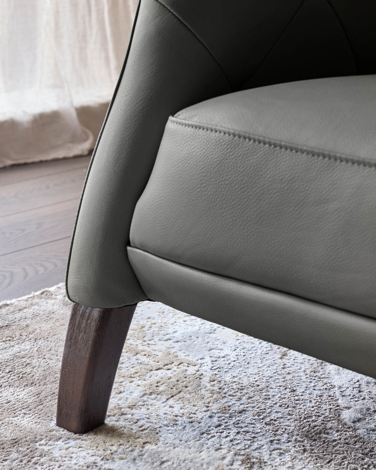 Close-up of a grey leather armchair showing the seat, arm, and a dark wood leg on a patterned rug.