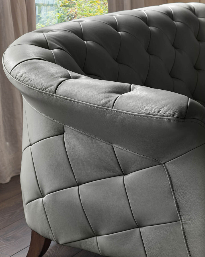 Close-up of a grey leather armchair with buttoned detailing and visible wooden leg. Subtle lighting from nearby window.