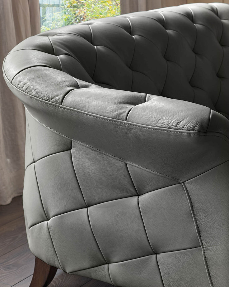 Close-up of a grey leather armchair with buttoned detailing and visible wooden leg. Subtle lighting from nearby window.