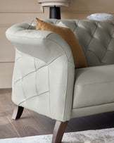 Close-up of a light grey leather sofa with a tufted back and arm, plus a brown velvet cushion and dark wood legs.
