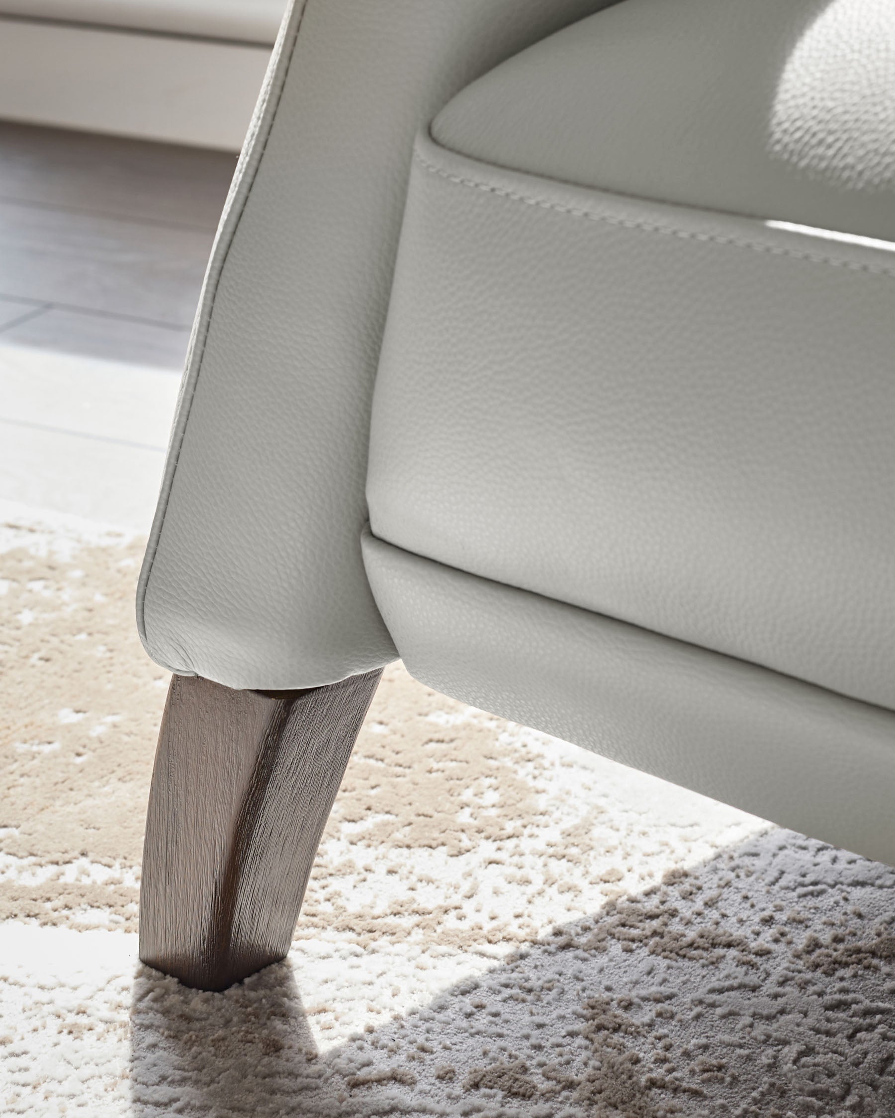 Close up of the light grey leather arm and dark wood leg of a stylish chair on a textured rug.