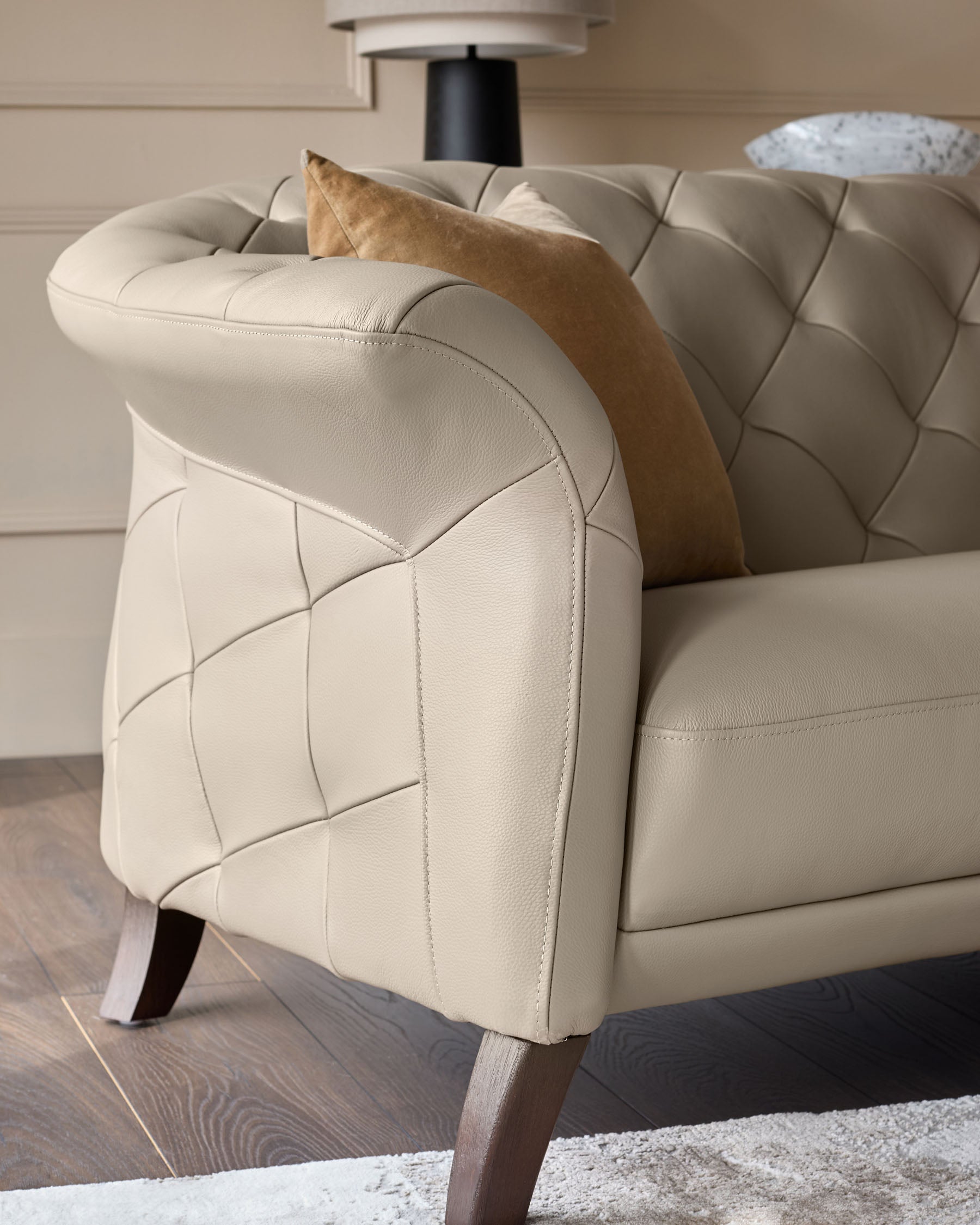 Close-up of a luxurious beige sofa with buttoned detailing and a brown cushion. Showing the arm, leg and part of the seat.