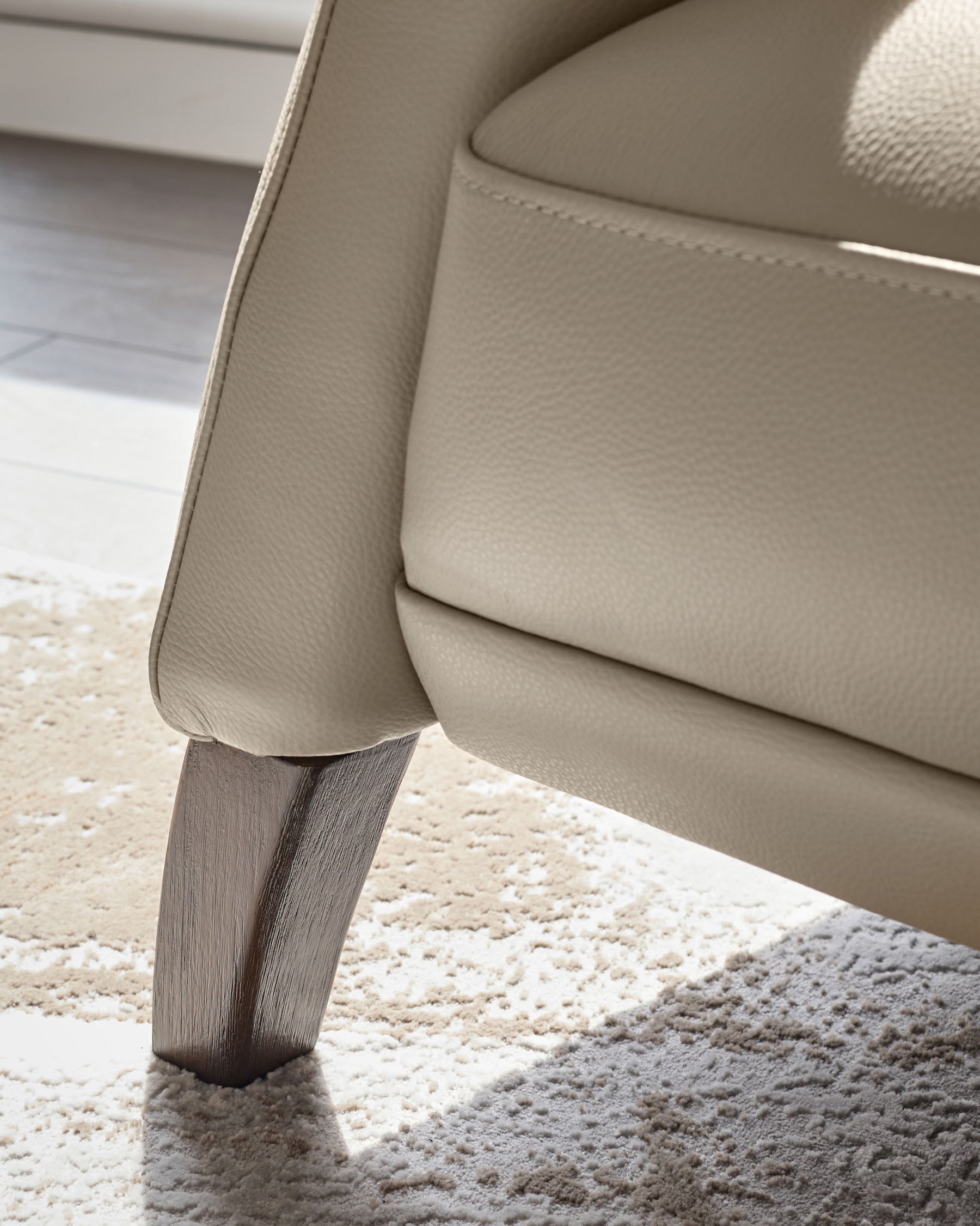 Close-up of a beige leather chair with a dark wooden leg, showcasing the texture of the chair and the patterned rug below.