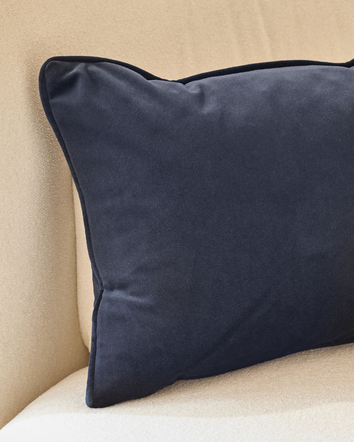 Navy rectangular cushion with piping detail, displayed on a cream-colored upholstered surface against a beige wall.