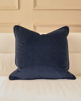 Navy blue velvet cushion with piping detail, displayed on a cream upholstered sofa against a paneled beige wall.