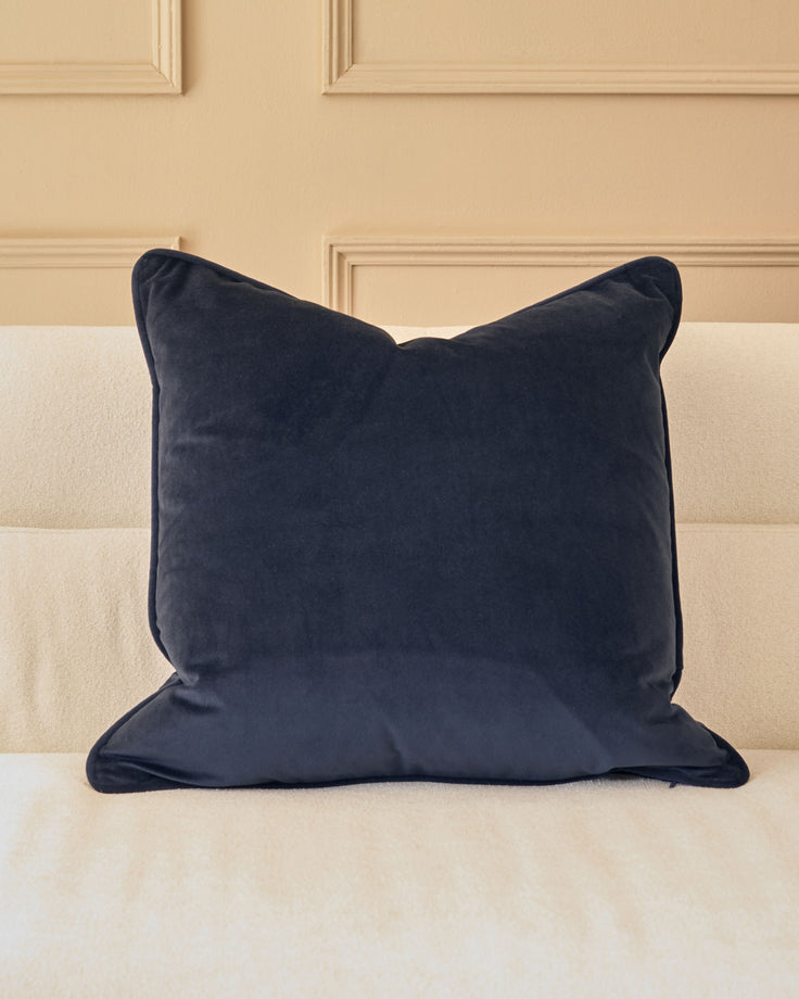 Navy blue velvet cushion with piping detail, displayed on a cream upholstered sofa against a paneled beige wall.