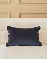 Navy blue rectangular velvet cushion with piped edges, displayed on a cream upholstered bed against a paneled headboard.