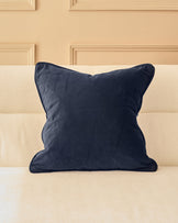 Navy velvet cushion with piping detail, displayed on a cream upholstered sofa against a beige paneled wall.