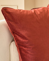 Luca medium cushion in toffee red velvet, displayed on a cream upholstered chair arm, showing soft texture and rich color.