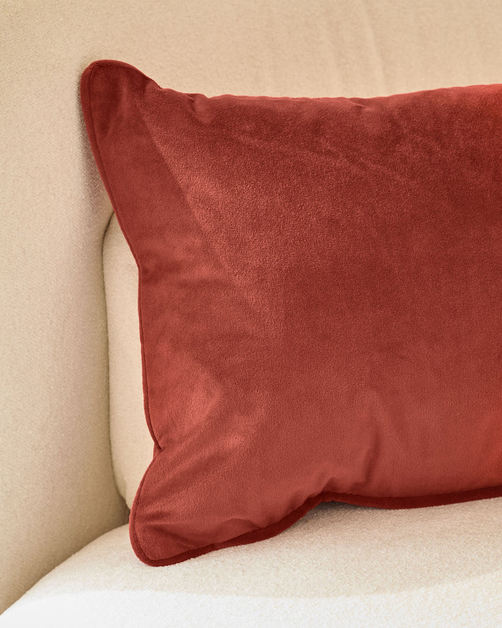 Luca rectangle cushion in toffee red, displayed on a cream sofa against a beige wall.