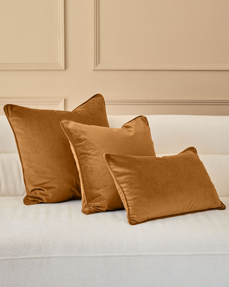 Luca Medium Caramel Cushion displayed on a white bed against a beige paneled wall, shown in a set of three.
