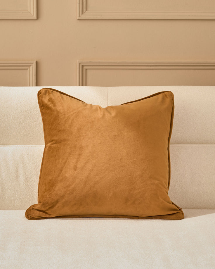 Caramel velvet cushion with piping on a cream upholstered bed against a beige paneled wall.