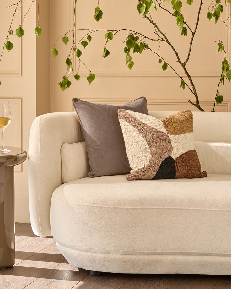 Lennon cushion in natural displayed on a cream curved armchair with geometric patterned pillows in a styled living room.