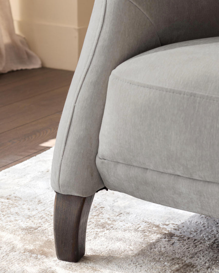 Close-up of a grey upholstered armchair with a curved arm and a dark wood leg on a light textured rug.