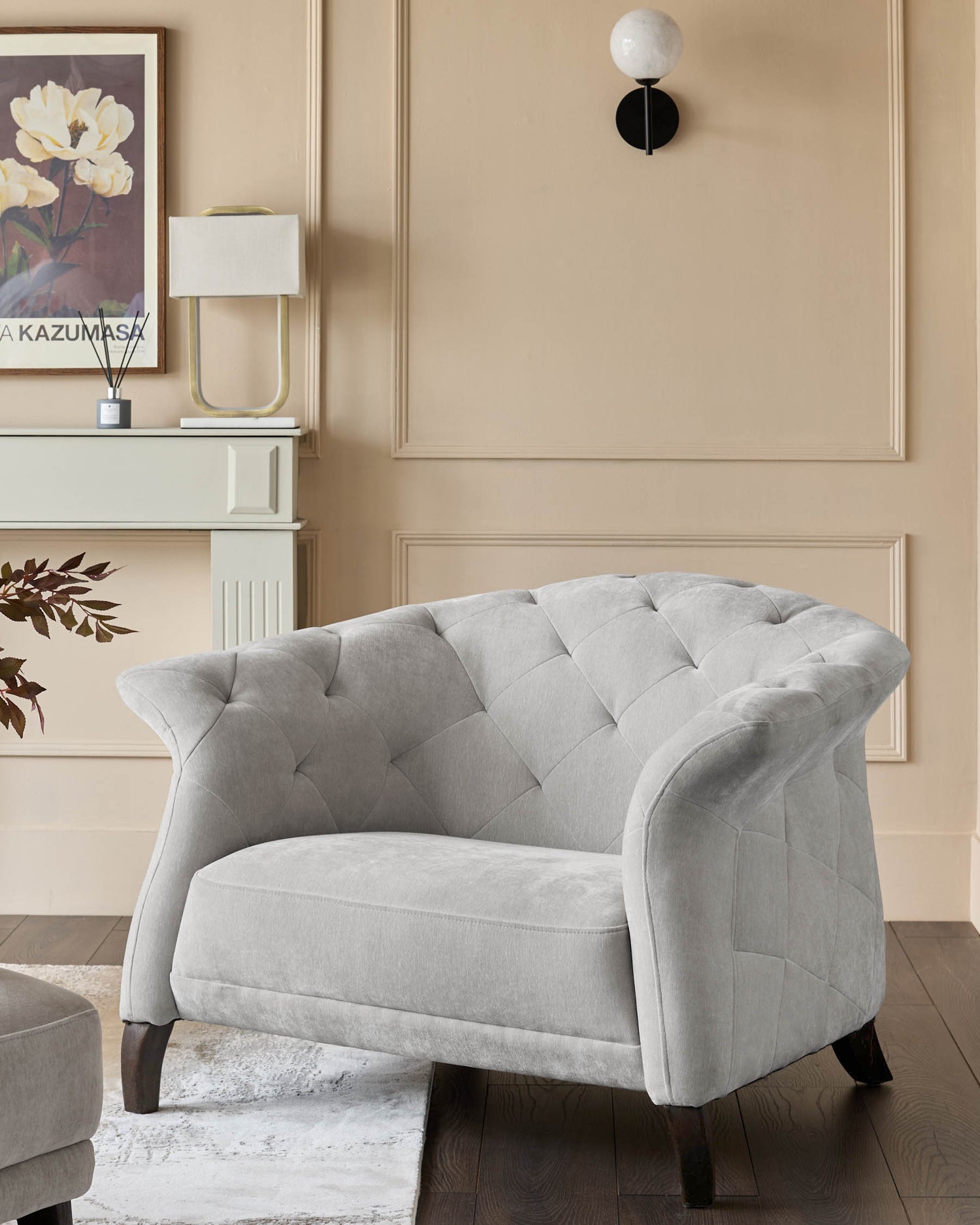 A grey, button-back armchair sits on a rug with a fireplace, lamp and wall sconce visible.
