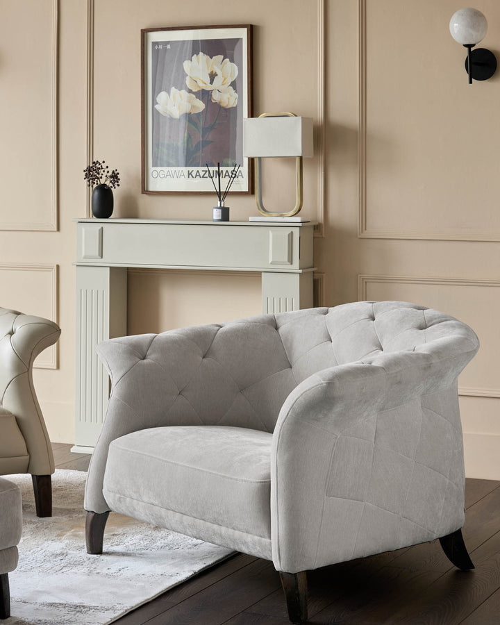 A light grey button-backed armchair sits on a patterned rug in a neutral living room. A cream fireplace is behind.