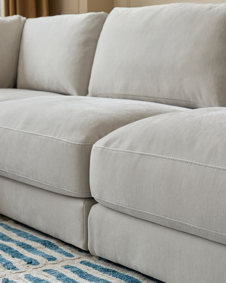 Lynetta small corner sofa in natural grey weave fabric, showing plush cushions and curved seating detail with patterned rug.