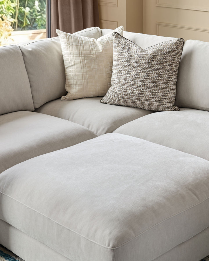 Lynetta small corner sofa with footstool in natural grey weave fabric, styled with decorative pillows in a bright room.