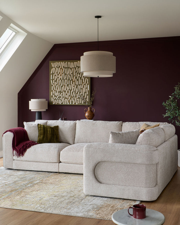 Lynetta small corner sofa in natural weave fabric, styled in a modern living room with deep burgundy walls and pendant lighting.