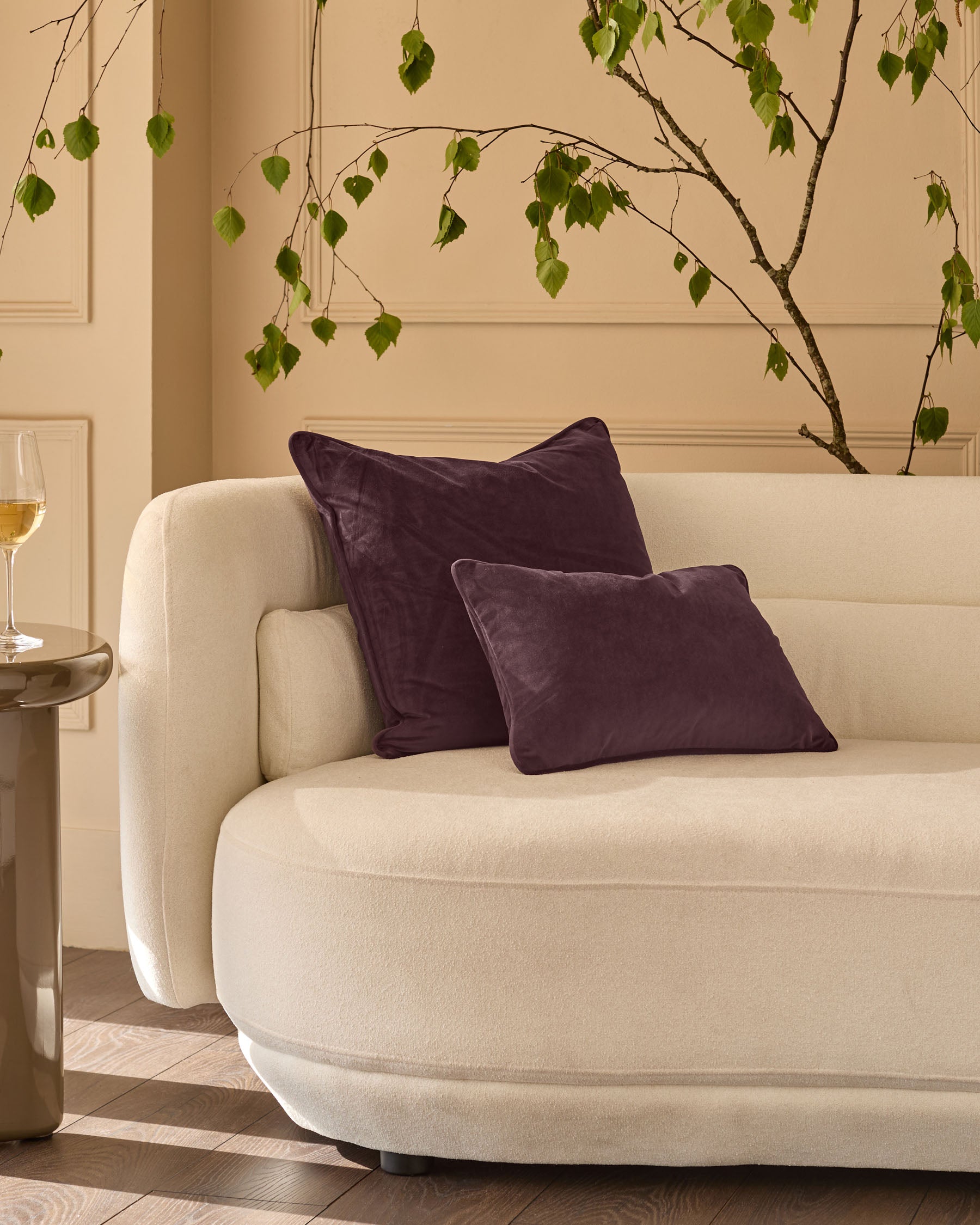 Luca cushion in mulberry, medium size, displayed on a cream curved sofa in a styled living room setting.