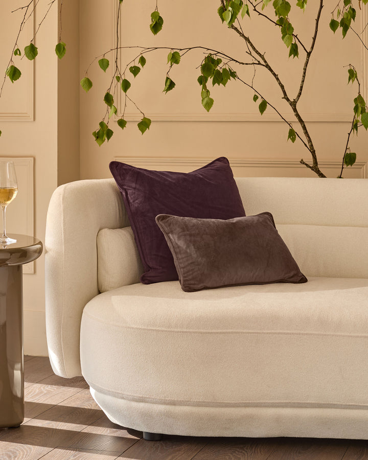 Large mulberry velvet cushion displayed on a cream armchair in a styled living room setting.