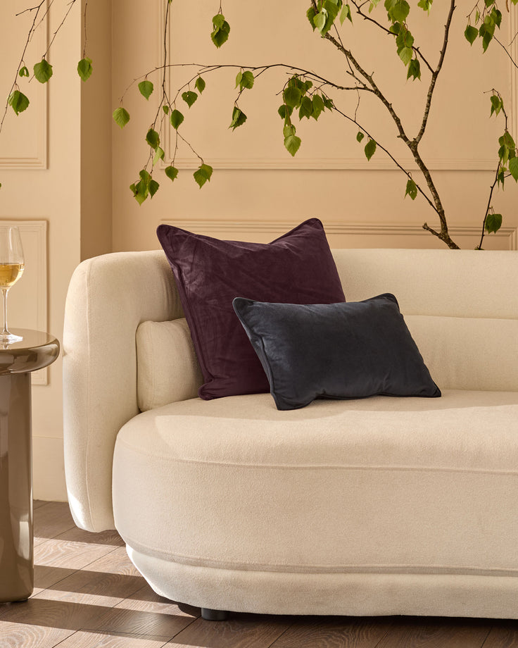 Large mulberry velvet cushion displayed on a cream curved armchair in a styled living room setting.