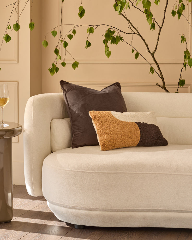 Large chocolate leather cushion displayed on a cream curved sofa in a styled living room setting.