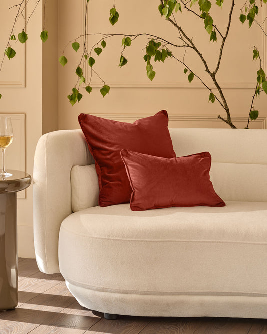 Luca medium cushion in toffee red, displayed on a cream curved chair with decorative green foliage above.