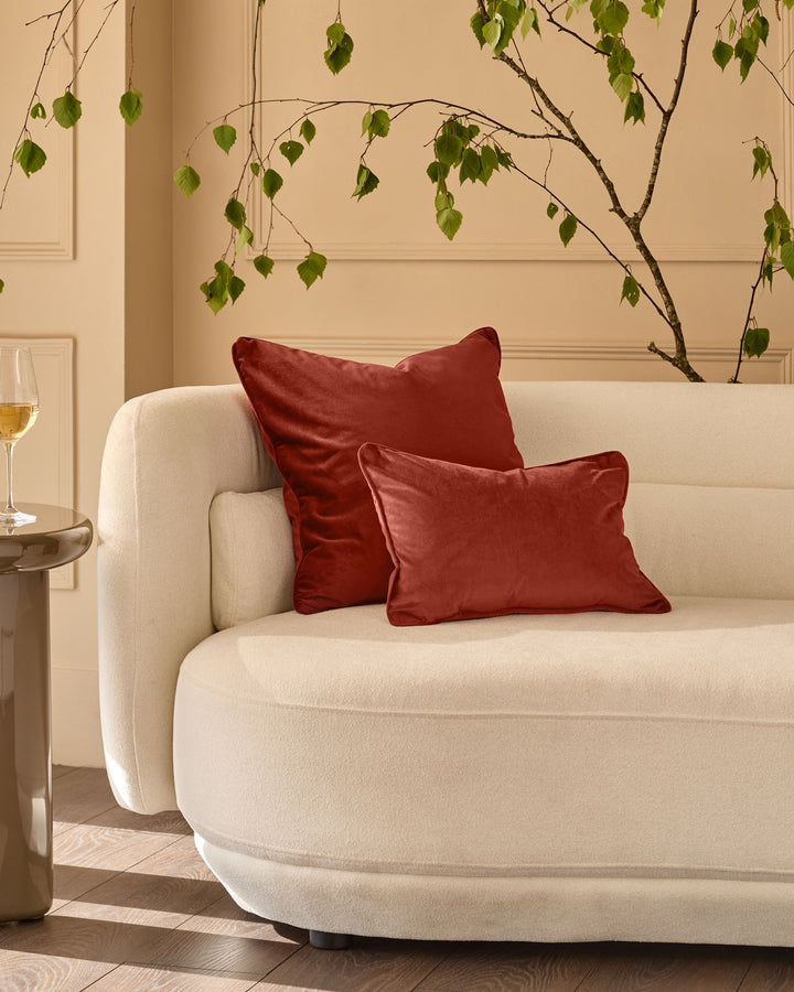 Toffee red rectangular cushion displayed on a cream curved armchair in a styled living room setting.