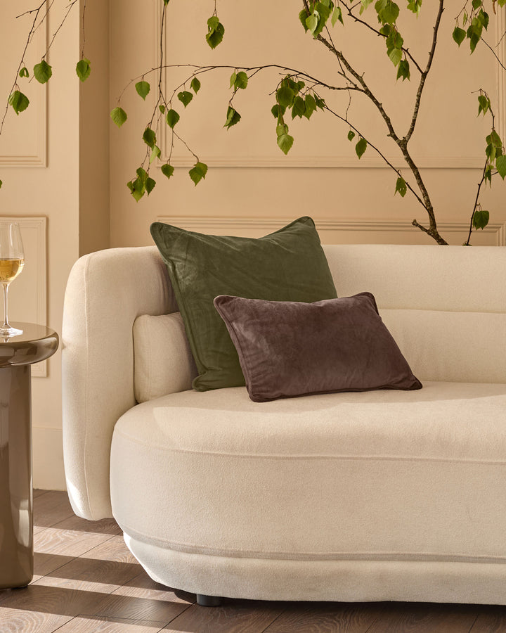 Luca medium olive green cushion displayed on a cream curved sofa alongside a burgundy cushion in a minimalist interior setting.