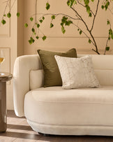 Ivory quilted square cushion displayed on a cream curved armchair in a styled living room setting.