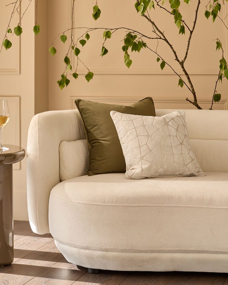 Ivory quilted square cushion displayed on a cream curved armchair in a styled living room setting.