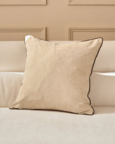 Madeira cushion in natural beige with brown piping, displayed on a cream sofa against a taupe paneled wall.