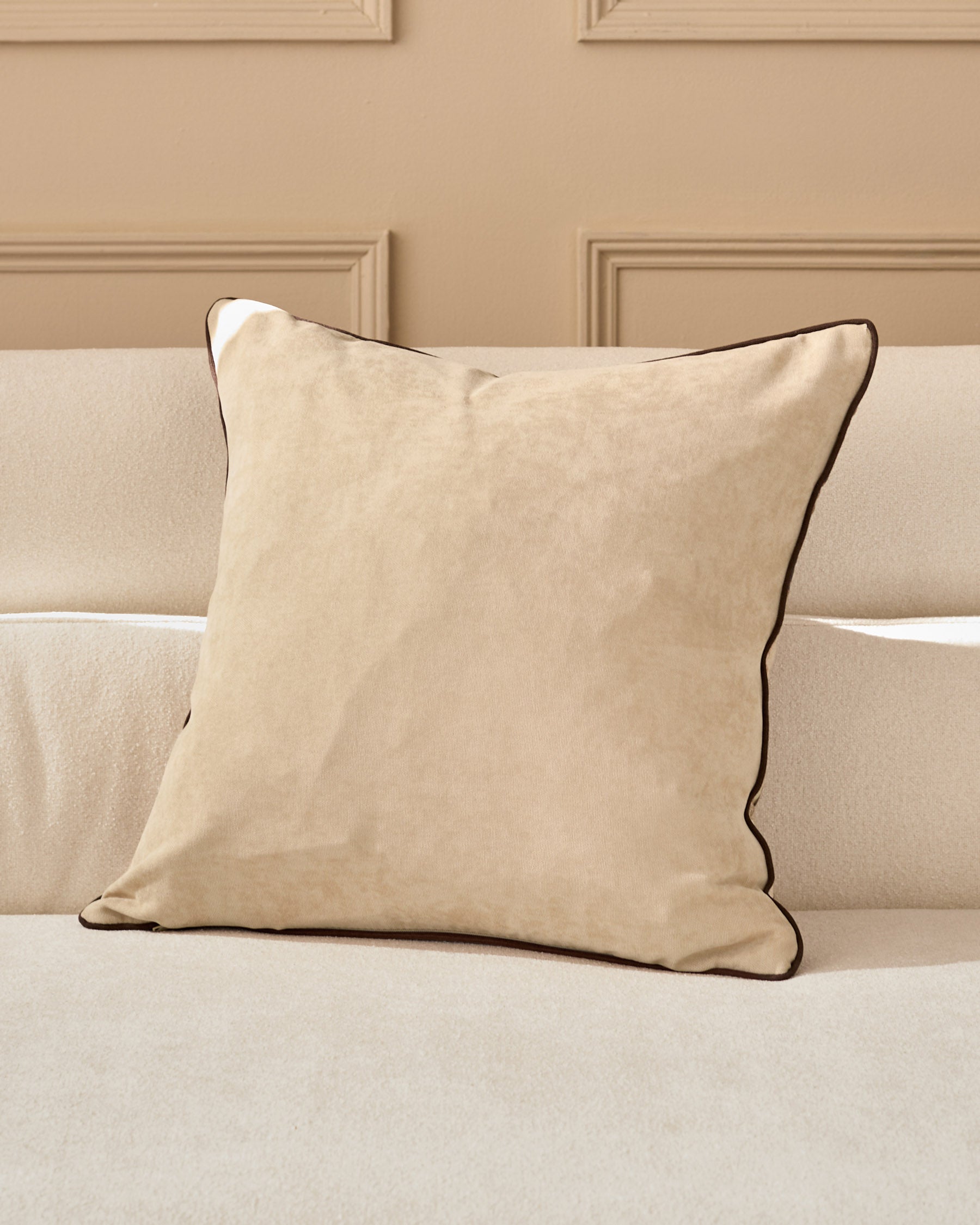 Madeira cushion in natural beige with brown piping, displayed on a cream sofa against a taupe paneled wall.