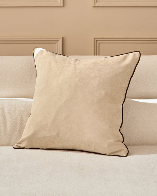 Madeira cushion in natural beige with brown piping, displayed on a cream sofa against a taupe paneled wall.
