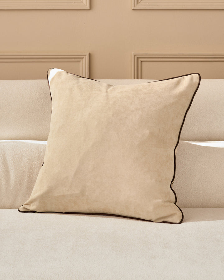 Madeira cushion in natural beige with brown piping, displayed on a cream sofa against a taupe paneled wall.