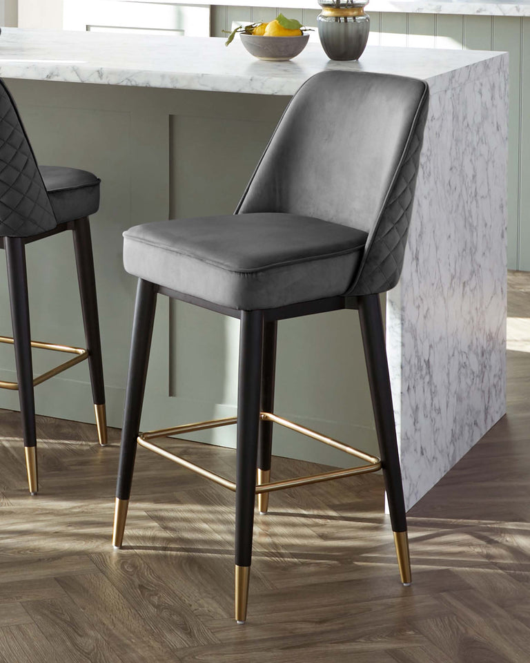 Elegant contemporary bar stool with dark grey upholstery and diamond tufting on the backrest. It features a sleek black frame with slender legs and metallic gold accents at the feet and footrest. The stool is shown next to a white marble countertop.