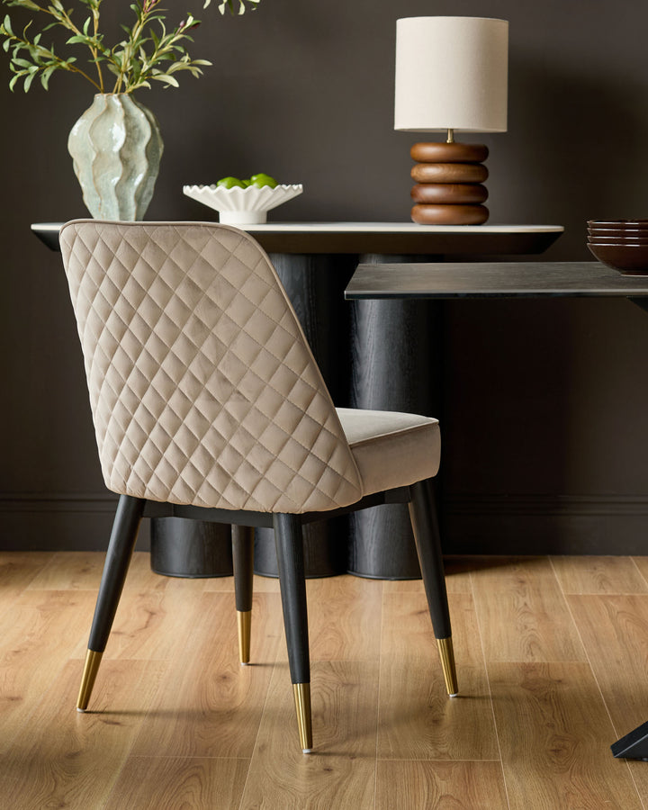 Mae velvet dining chair in champagne, shown at a dining table with quilted backrest and black legs with brass foot caps.