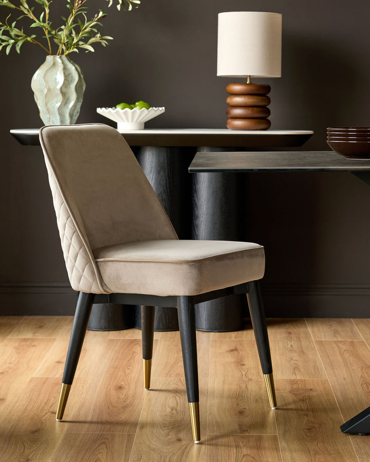 Mae champagne velvet dining chair with quilted backrest, black wooden legs with brass foot caps, shown at table.