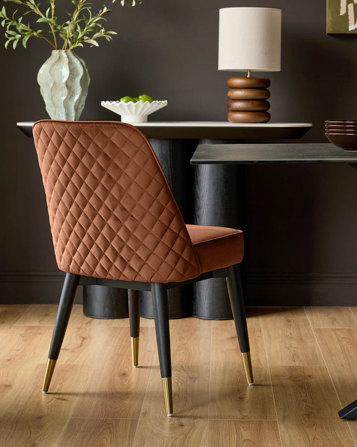 Mae velvet dining chair in rust with quilted backrest, black wooden legs, and brass foot caps, shown in styled room setting.