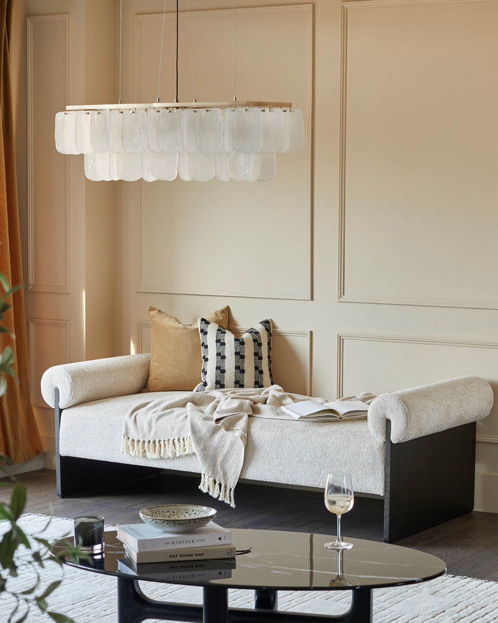 A stylish cream chaise longue with cushions, under a modern chandelier. A glass coffee table sits in front.