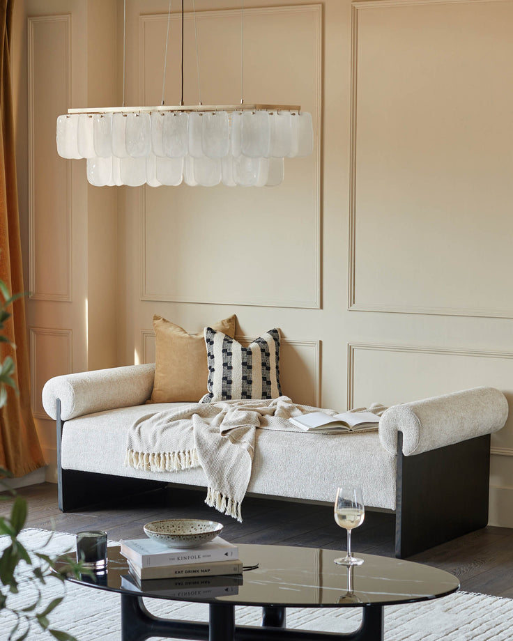 A stylish cream chaise longue with cushions, under a modern chandelier. A glass coffee table sits in front.