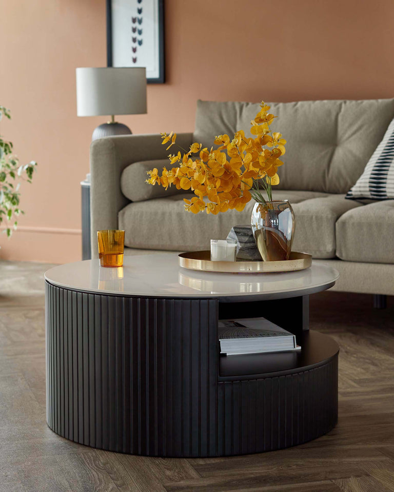 Modern round coffee table with a white top and a cylindrical base featuring vertical ribbed detailing. The base provides a storage space for books, and the tabletop has a reflective brass-coloured tray with a glass vase holding yellow orchids, adjacent to a golden glass and candle. Behind, a stylish beige sofa with a striped cushion completes the elegant setting.
