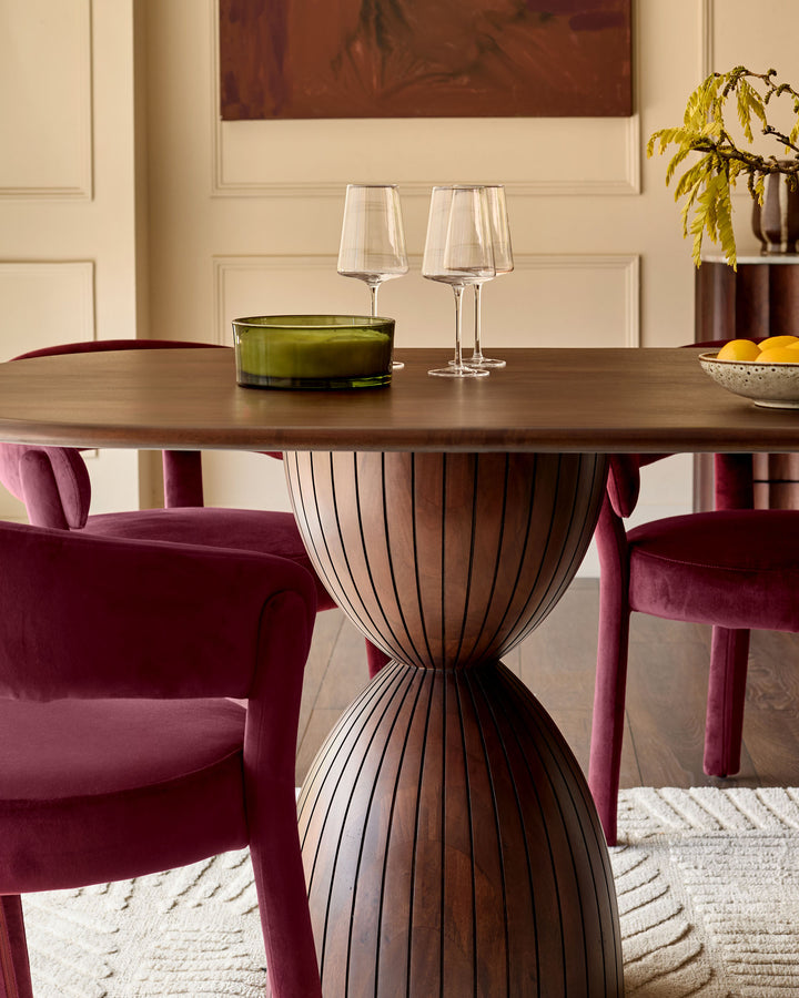 Malin dark mango wood dining table with sculptural hourglass-shaped base, styled with burgundy velvet chairs and place settings.