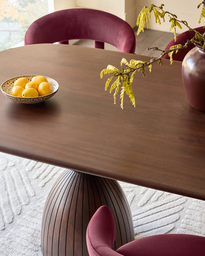 Malin round dining table in dark mango wood with pleated pedestal base, styled with burgundy chairs and yellow foliage.