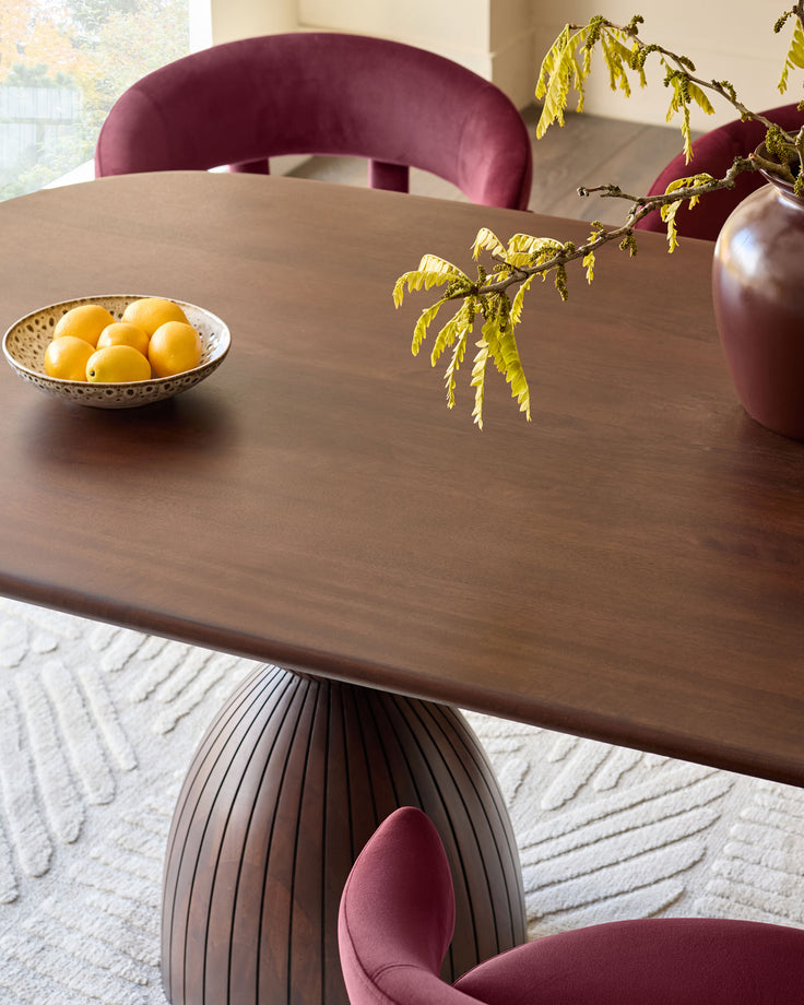 Malin round dining table in dark mango wood with pleated pedestal base, styled with burgundy chairs and yellow foliage.
