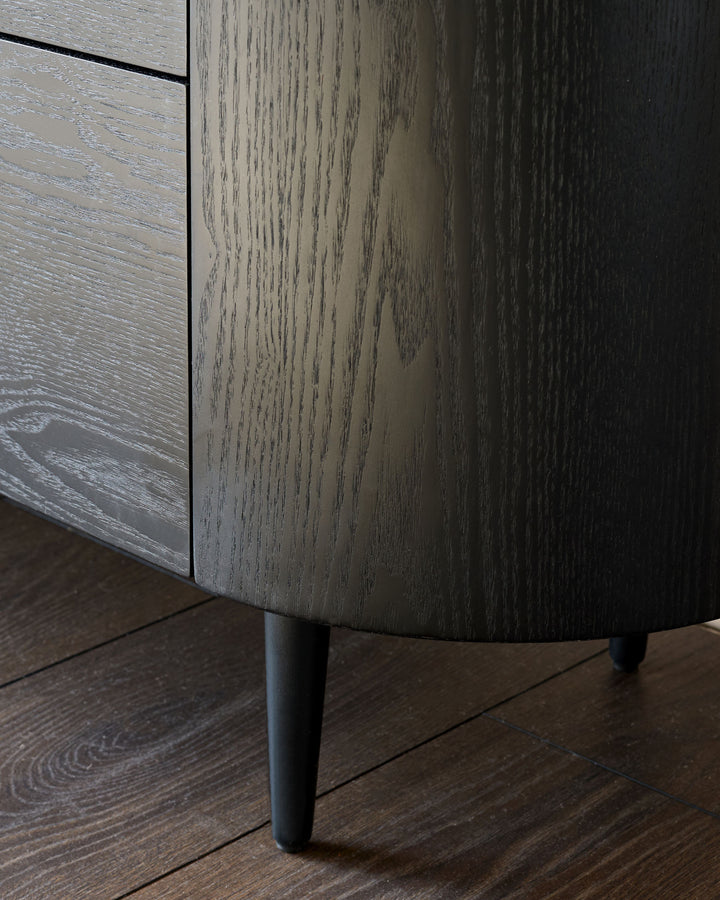 Manhattan 6-drawer chest in ceramic white with wood grain finish, angled side view showing tapered leg detail on dark wood floor.